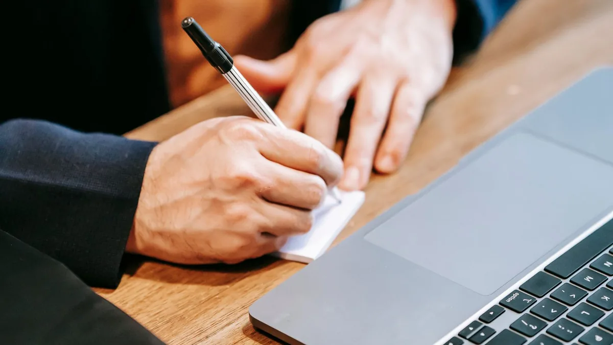 Hands writing notes beside a laptop on a wooden desk